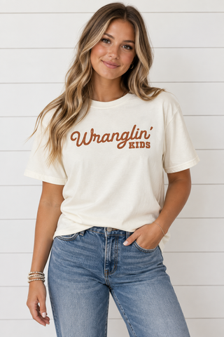 Woman wearing cream Wranglin’ Kids graphic tee with rust lettering and blue jeans