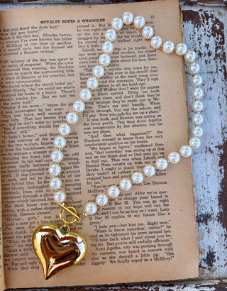 White pearl-style beaded necklace with gold-tone spacers, a toggle clasp, and a large gold-tone heart pendant, displayed on a vintage book page.