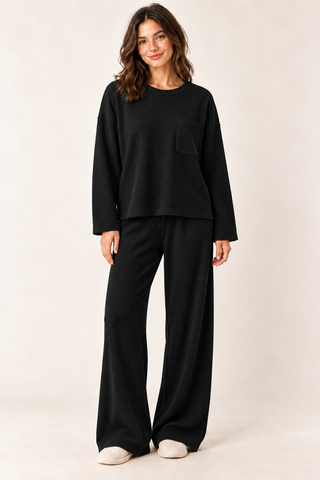 Model wearing a black waffle knit lounge set with a long\-sleeve crewneck top, chest pocket, and matching wide\-leg pants\.