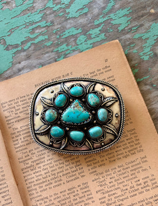 Antique silver-tone belt buckle with a large turquoise-colored center stone, eight surrounding stones, leaf accents, and a twisted rope border.