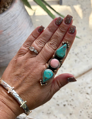 A close-up of a hand wearing a silver-toned statement ring with two large turquoise teardrop stones and one round pink stone, along with a silver bangle bracelet.