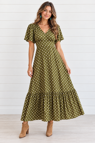 Model wearing an olive green polka dot maxi dress with flutter sleeves and a ruffled hem, styled with tan ankle boots.