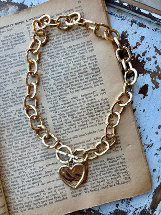 Gold-plated necklace with chunky textured oval links, a front toggle clasp, and a heart pendant, displayed on an open book.
