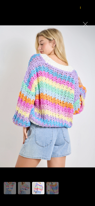 A woman wearing a colorful crochet knit sweater with pastel rainbow stripes and light blue denim shorts, shown from the back against a plain light gray background.