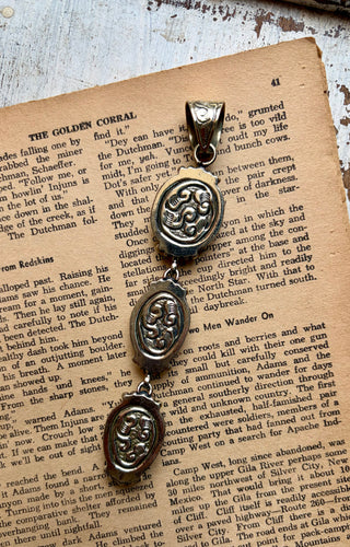 A silver-toned metal pendant with an engraved floral pattern, shown hanging against a background of vintage book pages.