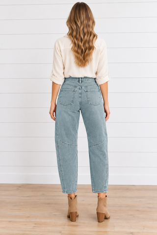 Model wearing washed teal barrel jeans with curved seams and knee darts, a cream blouse, turquoise necklace, and tan ankle boots.