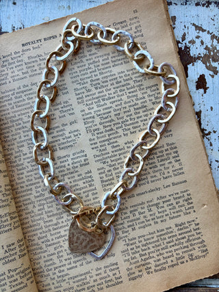 Gold- and silver-tone chunky chain necklace with a front toggle clasp and overlapping solid and open heart pendants, displayed on a book.