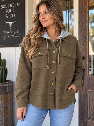 Woman wearing the Timberline olive-brown plaid hooded shacket with gray hood, brass snap buttons, and front chest pockets.