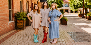 Three women standing on a brick-paved outdoor walkway in front of a brick building, wearing long dresses in beige, gray, and blue with boots. A black sign above them reads 'Simplifi'.
