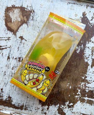 Translucent yellow jumbo banana squishy with colorful fruit-shaped pieces inside, displayed in a clear box on distressed white wood.