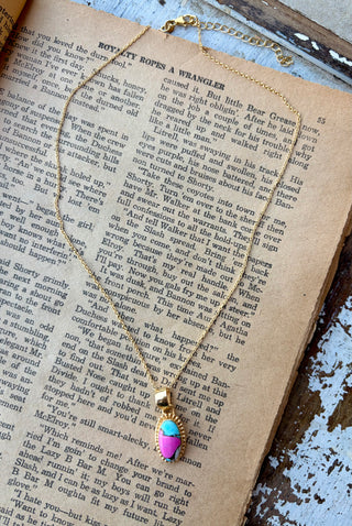 Oval pink and turquoise-blue Cotton Candy stone pendant with a beaded gold border on a gold-plated sterling silver chain, displayed on an open book.