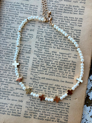 White pearl-look beaded necklace with gold-tone cross accents, small spacers, and an extender chain, displayed on an open book.