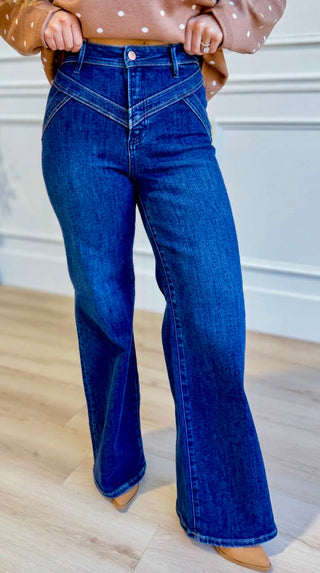 Model wearing Risen dark-wash high-rise wide-leg jeans with angled front yoke seams, an ivory tank, and tan boots.