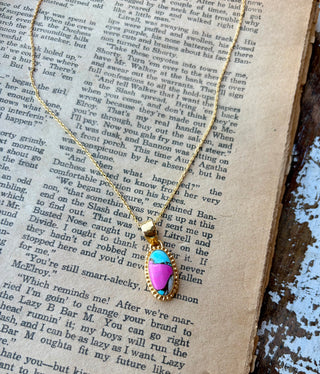 Oval pink and turquoise-blue Cotton Candy stone pendant with a beaded gold border on a gold-plated sterling silver chain, displayed on an open book.