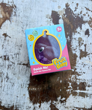 Purple Fruit Baby squishy in blue and pink packaging labeled “Squish Me!” and “3+,” displayed on distressed white wood.