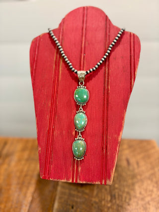 Three-stone turquoise pendant necklace with silver-tone detailing and beaded necklace, displayed on a rustic red wooden jewelry stand.