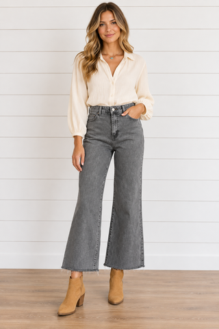 Model wearing ash black high-rise wide-leg cropped jeans with raw hems, a cream blouse, turquoise necklace, and tan ankle boots.