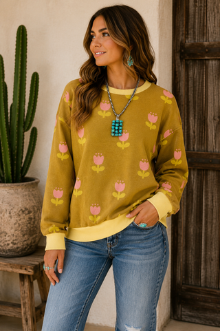 A woman wearing a mustard yellow crewneck sweatshirt with a pink and yellow floral pattern, paired with blue jeans, standing in front of a rustic wooden door and a potted cactus.