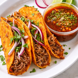 Three birria beef tacos topped with red onion and cilantro, served with consommé for dipping, lime wedges, and sliced radishes.