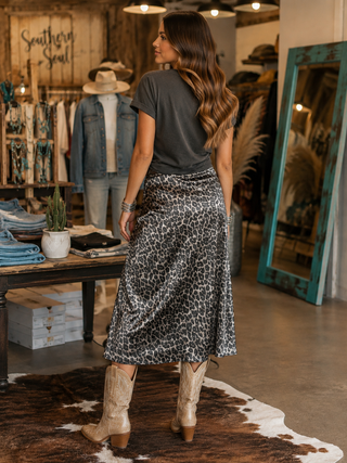 A woman stands in a rustic boutique wearing a dark gray t-shirt and a black and white leopard print midi skirt, paired with beige cowboy boots. The background features a wooden sign reading 'Southern Soul', a mannequin in a denim jacket, and a large teal-framed mirror.