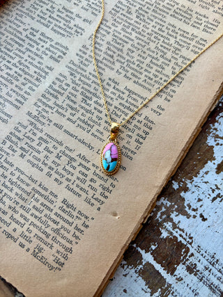 Oval pink and turquoise-blue Cotton Candy stone pendant with a beaded gold border on a gold-plated sterling silver chain, displayed on an open book.
