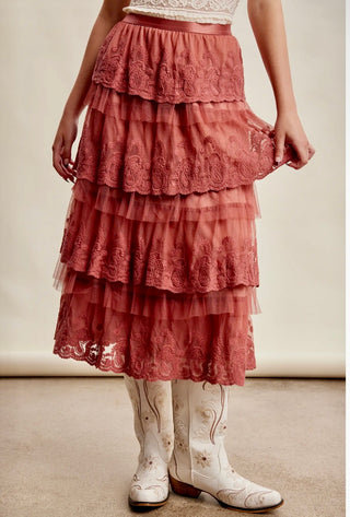 Model wearing a rust-colored tiered lace midi skirt with ruffle detailing, styled with an ivory lace top and white embroidered western cowboy boots.