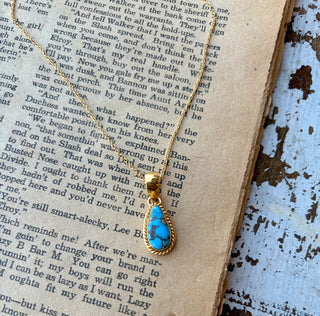 Blue Golden Hills turquoise teardrop pendant with dark veining and a gold rope-style border on a gold-plated sterling silver chain, displayed on an open book.