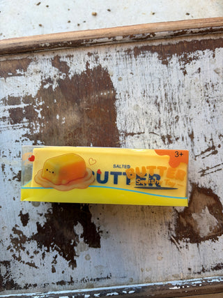 Butter squishy toy in yellow packaging featuring a smiling butter illustration, displayed on a distressed wood tray.