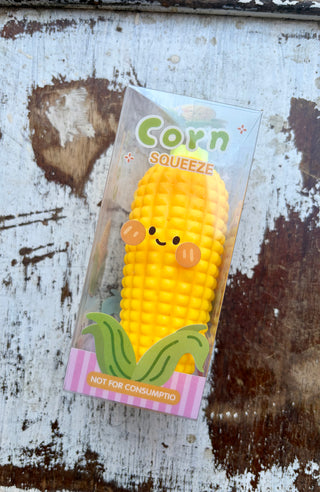 Yellow corn-shaped squishy with textured kernels in a clear box labeled “Corn Squeeze,” with a smiling face graphic and green husk illustrations.
