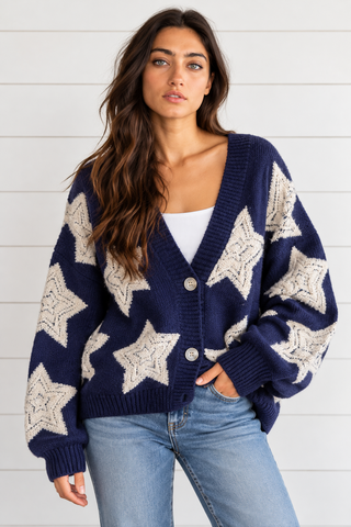 Model wearing an oversized navy button\-front cardigan with cream stars, ribbed trim, and long sleeves, styled with denim and boots\.