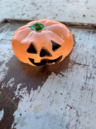 Orange pumpkin squishy with a black smiling jack-o’-lantern face and green stem on a distressed wood surface.