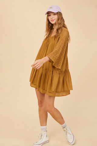 A woman modeling a loose, tiered mustard yellow dress with three-quarter crochet sleeves, paired with a lavender baseball cap, white socks, and white high-top sneakers, set against a solid beige studio background.
