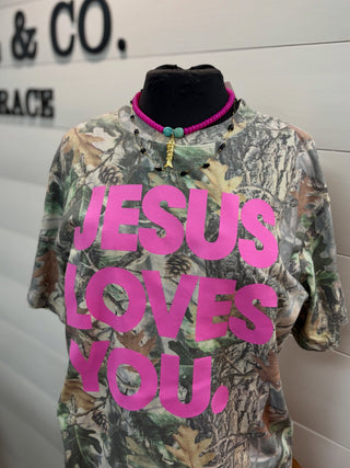 A camouflage graphic t-shirt displayed on a black mannequin torso, featuring the text 'JESUS LOVES YOU,' in bold hot pink. The shirt has a woodland camo pattern with green, brown, and gray tones.